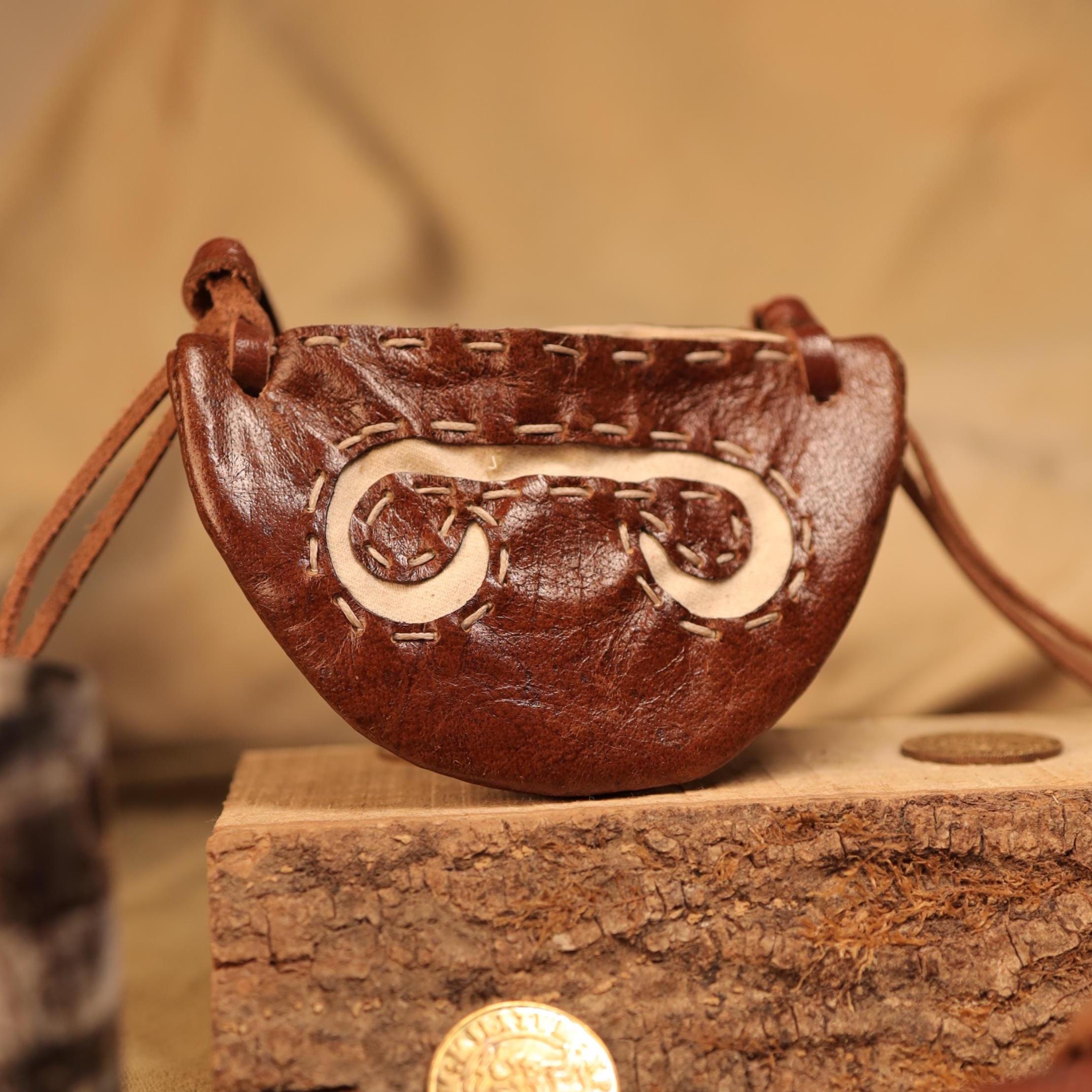 The Gokstad Wallet Viking Coin Pouch, a small handcrafted brown leather pouch with white stitching and a leather strap, shaped like a semicircle, sits on wood with a coin nearby. It has a rustic, artisanal look.