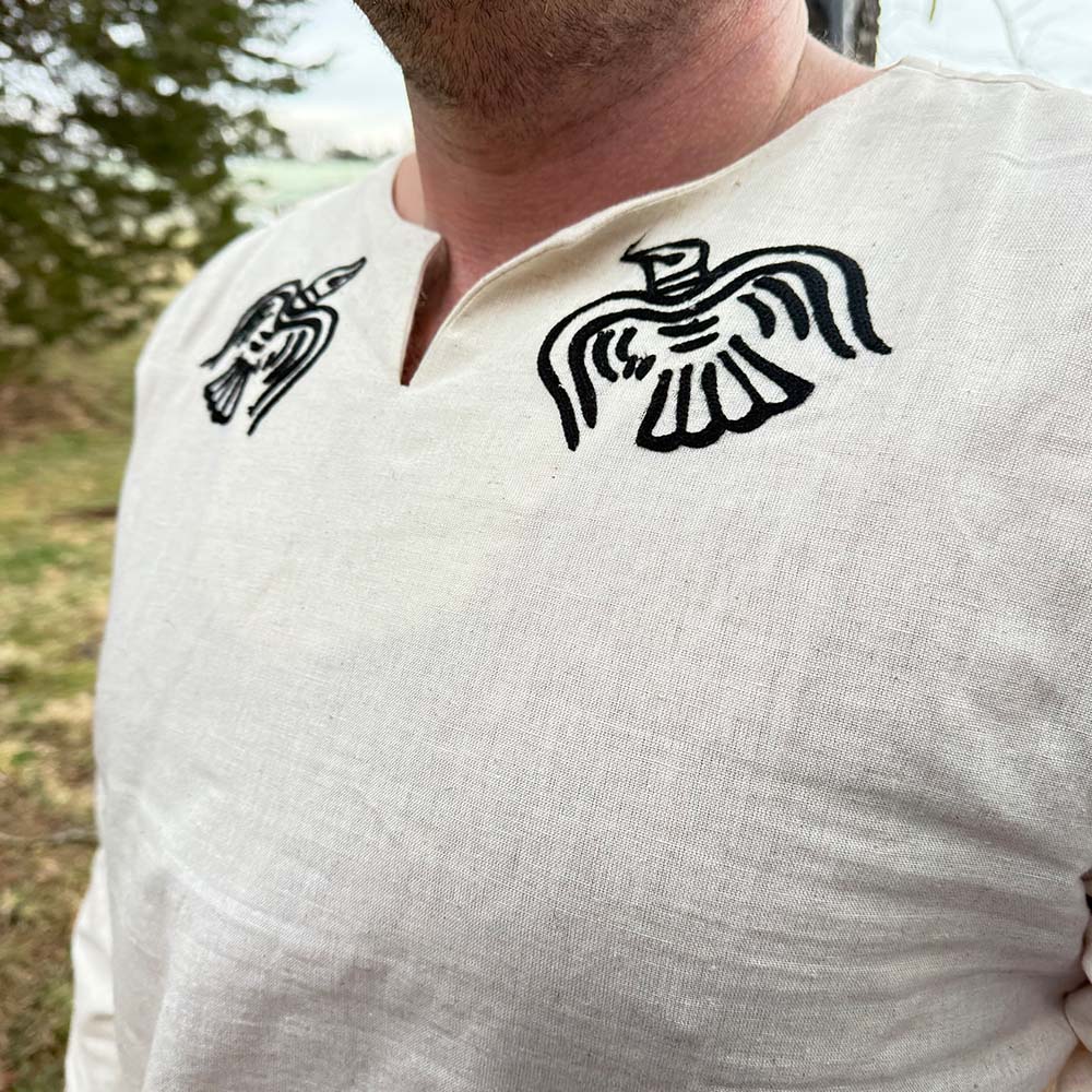 A person wearing the Odin's Raven Tunic- Men's, a cream Viking tunic with black raven designs on the shoulders and a notched neckline, stands outdoors with trees and grass in the background.
