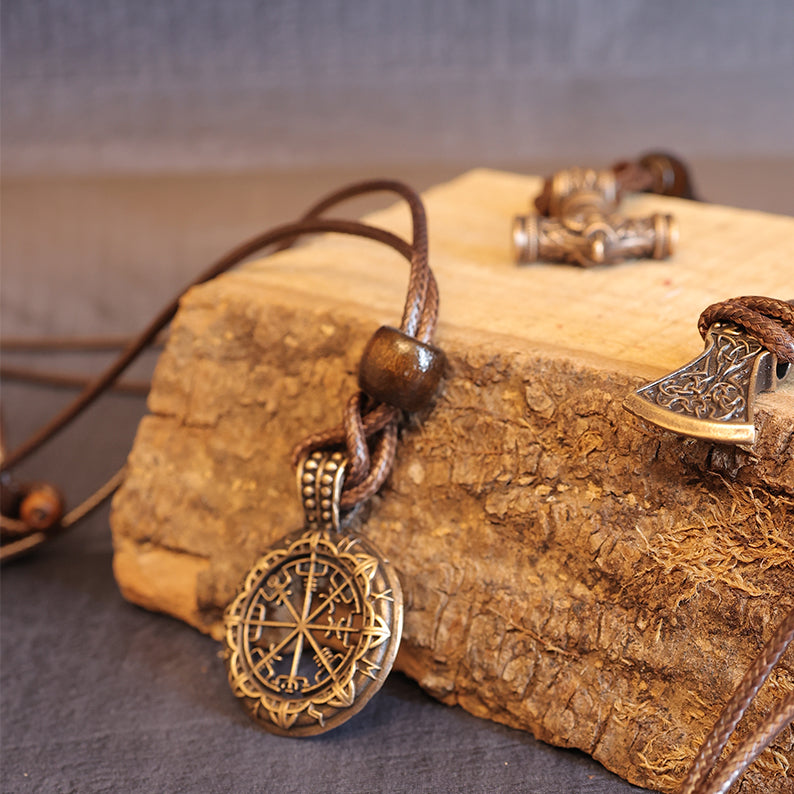 The Nomad's Compass Pendant, an ornate axe-shaped Viking necklace with intricate designs and a brown leather cord, rests on rustic wood—perfect as travel jewelry or a cherished Norse talisman.