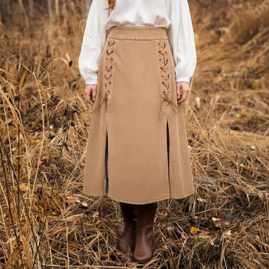 A person stands outdoors in a field of dry grass, wearing a white long-sleeve blouse, brown boots, and the Medieval Skirt- Ladies—a tan skirt featuring lace-up details and front slits.