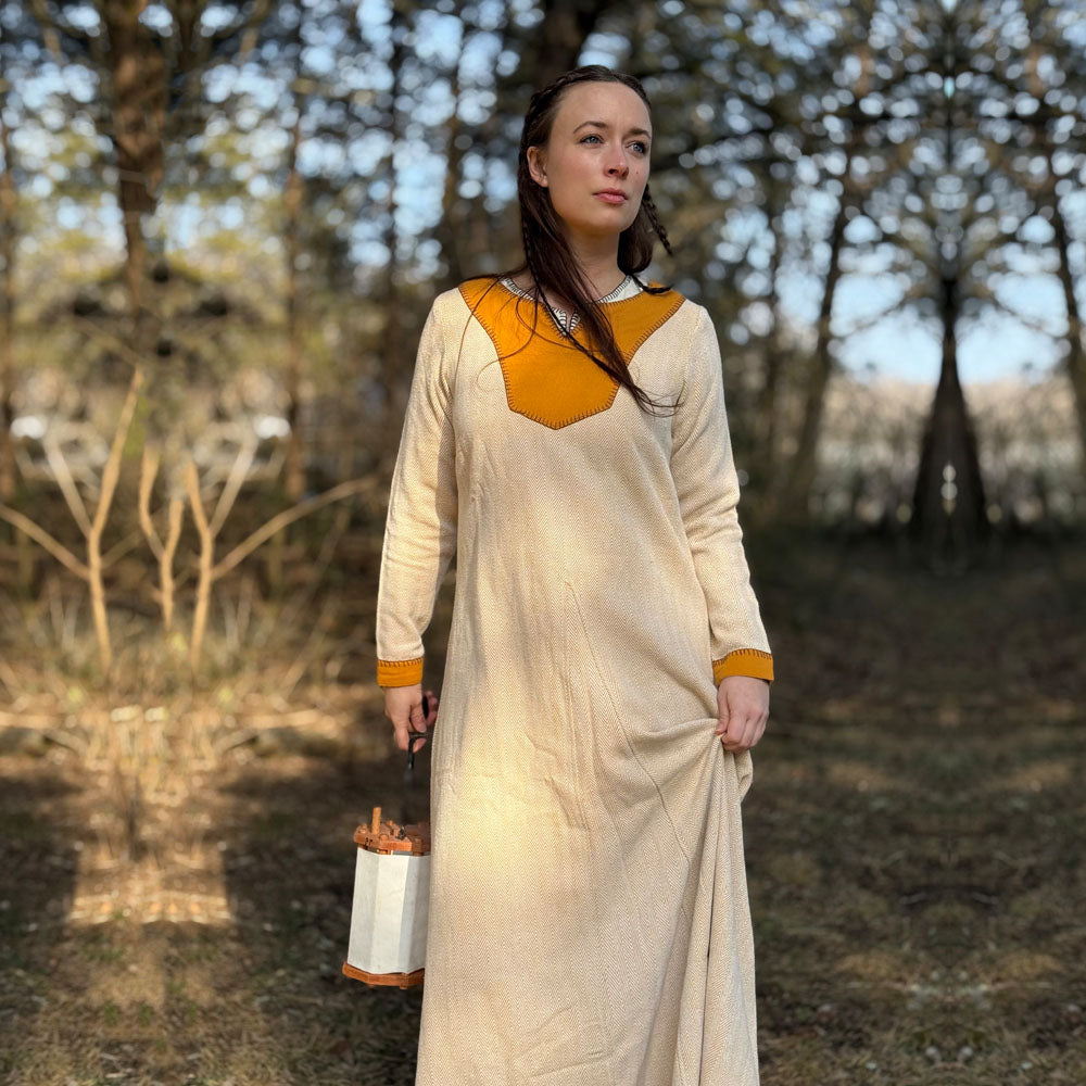 A woman wearing the Nordic Runeweave Viking Dress-Ladies, a long cream-colored gown with orange accents, stands outdoors under trees holding a lantern, sunlight highlighting the detailed Norse craftsmanship of her attire.