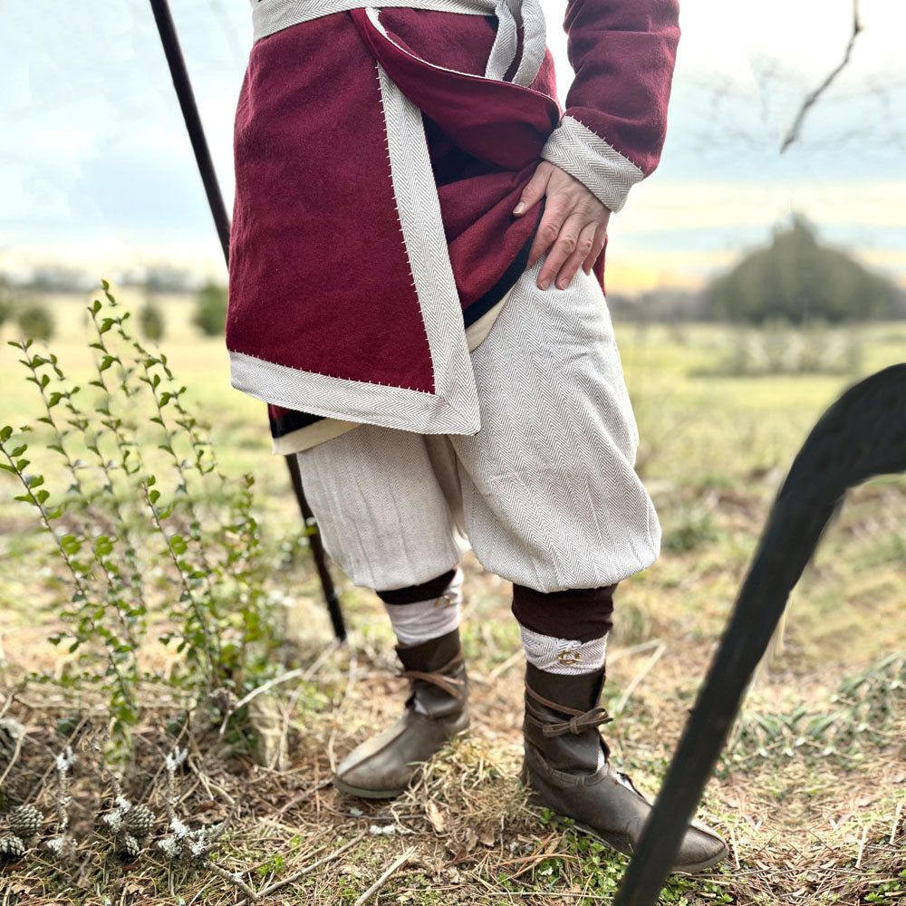 A person wearing Nordic Nomad Viking Pants stands outdoors on grass in historical clothing, with brown boots and leg wraps. The upper body is out of frame; the background has plants and a soft-focus landscape.