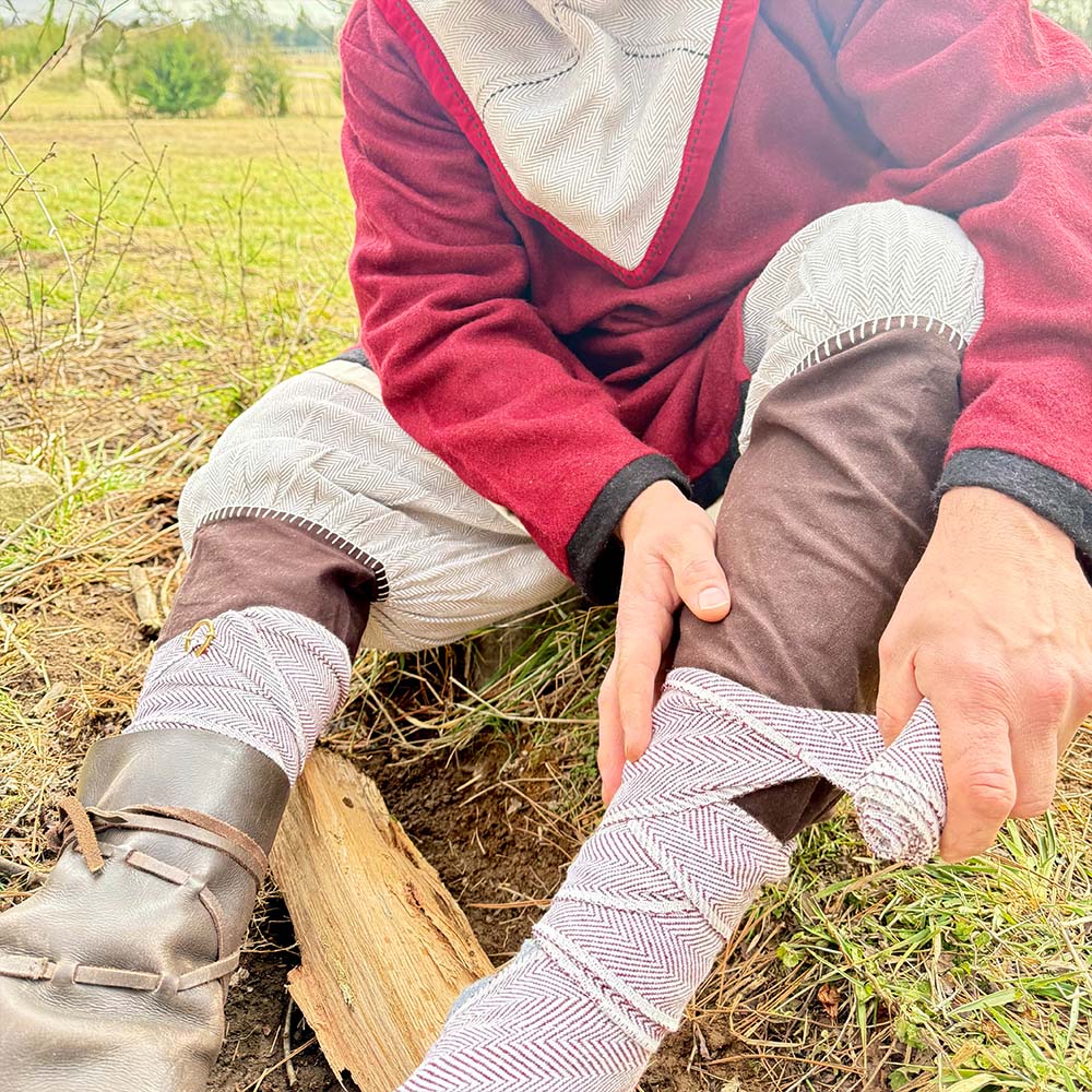 Outdoors on green grass, a person in historical attire wraps Herringbone Viking Leg Wraps (Winingas) over brown trousers, with leather shoes visible.