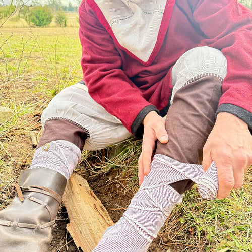 Outdoors on green grass, a person in historical attire wraps Herringbone Viking Leg Wraps (Winingas) over brown trousers, with leather shoes visible.