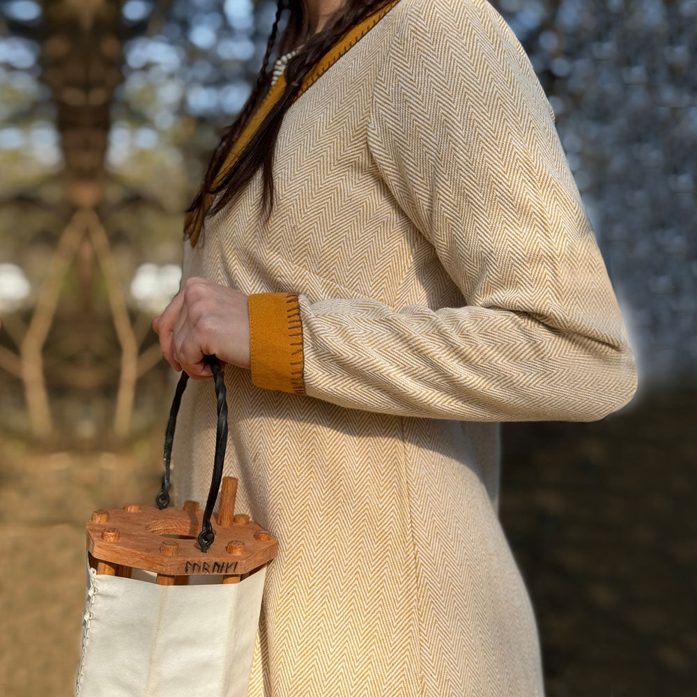 Wearing the Nordic Runeweave Viking Dress-Ladies in beige herringbone with mustard trim, a woman holds a small white and wooden bucket-style bag outdoors, trees softly blurred behind her—a modern tribute to Norse style.