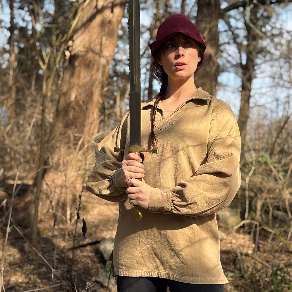 Wearing the Warrior Shirt – Medieval Strength & Style- Unisex and a red cap, a person stands in a sunlit forest, gripping a large sword upright with both hands, their expression serious as they look ahead.
