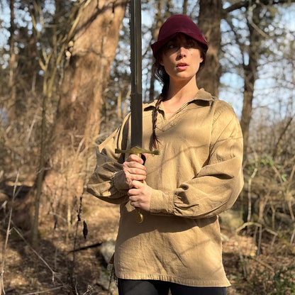 Wearing the Warrior Shirt – Medieval Strength & Style- Unisex and a red cap, a person stands in a sunlit forest, gripping a large sword upright with both hands, their expression serious as they look ahead.