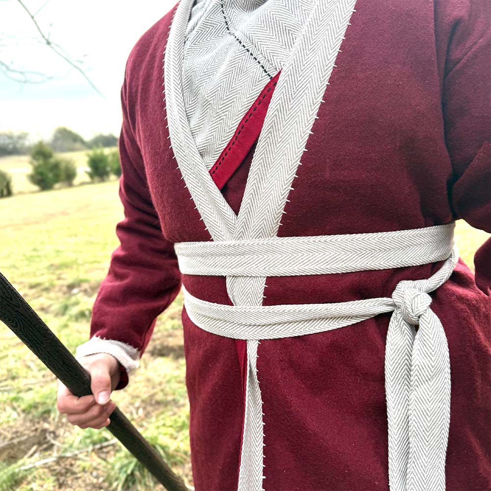 A person stands in a grassy field wearing the Viking Wool Coat- Men's in maroon with white trim and a white sash at the waist, holding a wooden staff.