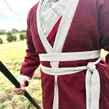 A person stands in a grassy field wearing the Viking Wool Coat- Men's in maroon with white trim and a white sash at the waist, holding a wooden staff.