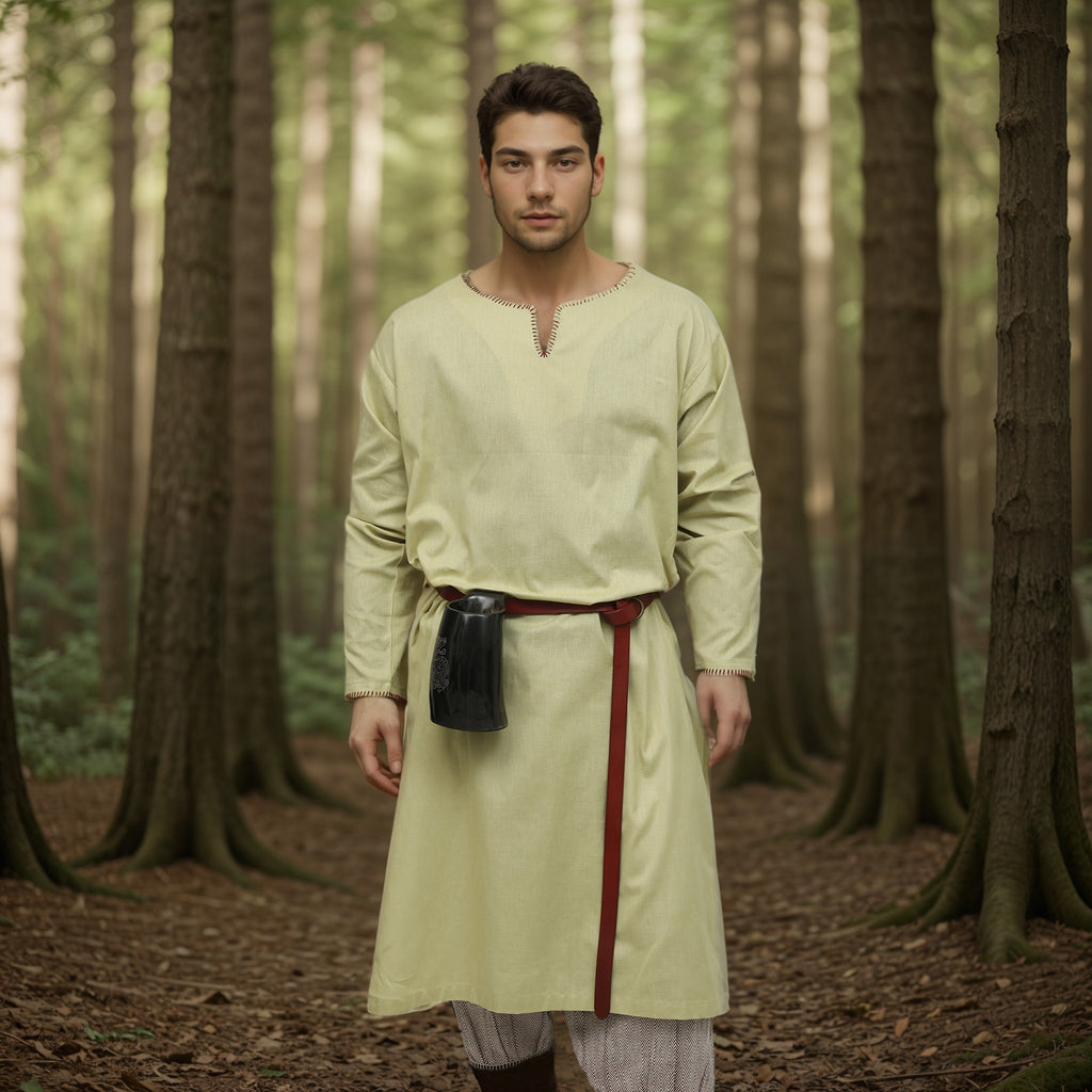 A man stands in a sunlit forest wearing the Viking Thorndale Tunic- Men's with a red belt and dark pouch, surrounded by tall trees—ideal for medieval cosplay or historical reenactment.