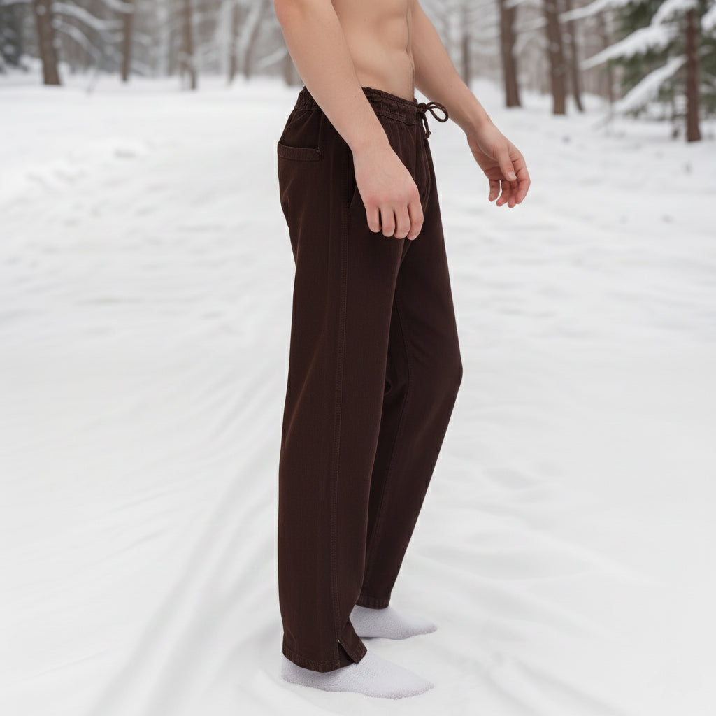 A person stands barefoot in the snow amid snowy trees, wearing Warrior Pants- Men's and white socks. Their upper body is bare, and their face is not visible.