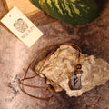 The Viking Rune Pendant Fehu with a striking rune design hangs from a brown leather cord, resting on textured rock. A Viking Alley logo tag and a green leaf are visible in the background.