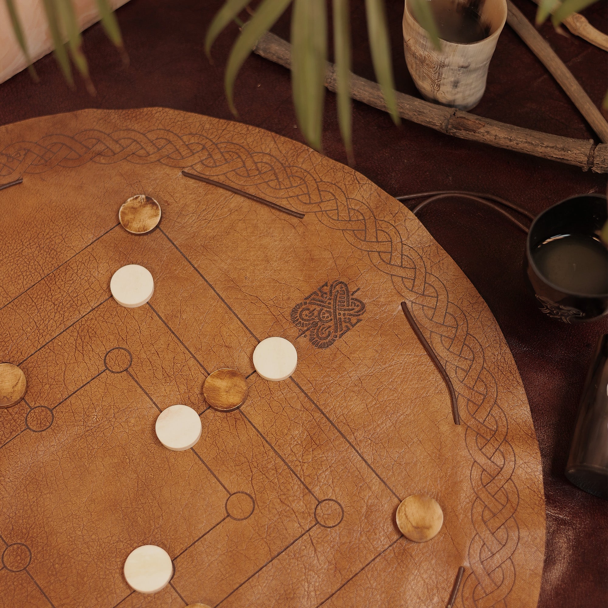 The Nine Men's Morris game features a leather board with a Celtic knot design and comes with white and brown pieces for classic two-player play. Also shown are a cup of dark liquid, wooden items, and part of a plant leaf nearby.