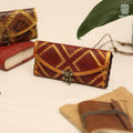 The Skald's Satchel is a handcrafted leather wallet featuring red and brown geometric Norse-inspired patterns and a metal clasp, displayed on a cream surface beside a notebook, burlap-wrapped book, and green leaf.