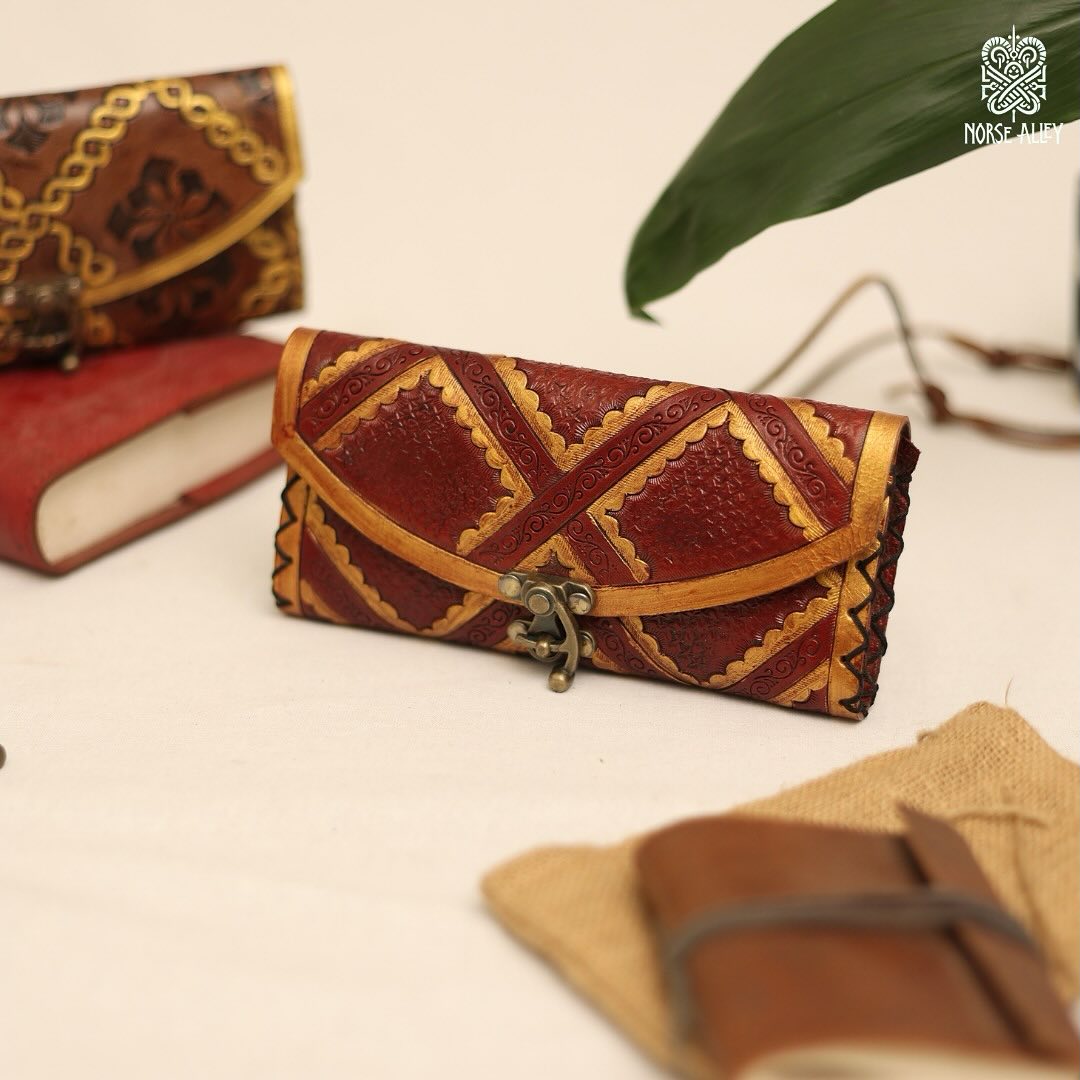 The Skald's Satchel is a handcrafted leather wallet featuring red and brown geometric Norse-inspired patterns and a metal clasp, displayed on a cream surface beside a notebook, burlap-wrapped book, and green leaf.