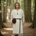 A young person in the Viking Thorndale Tunic–Men’s, with long blond hair and a belt and pouch, stands on a sunlit forest path among tall trees, embodying the feel of historical reenactment.