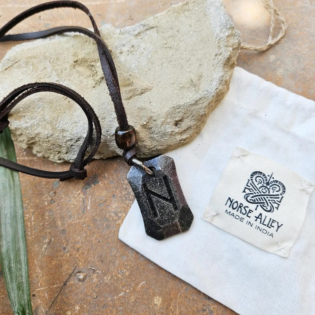 The Viking Rune Pendant Hagalaz features a black pendant engraved with the Hagalaz rune on a brown cord, displayed with a rough stone and a white Norse Alley pouch labeled Made in India, reflecting Norse mythology.