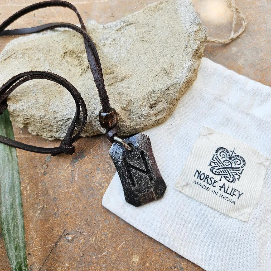 The Viking Rune Pendant Hagalaz features a black pendant engraved with the Hagalaz rune on a brown cord, displayed with a rough stone and a white Norse Alley pouch labeled Made in India, reflecting Norse mythology.