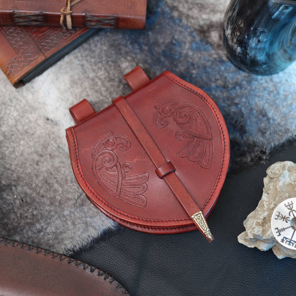 The "Huginn and Muninn" Viking Bag, featuring intricate embossing and a decorated clasp, rests on fur alongside a black leather item, a Norse mythology-inspired rune stone, and a journal.