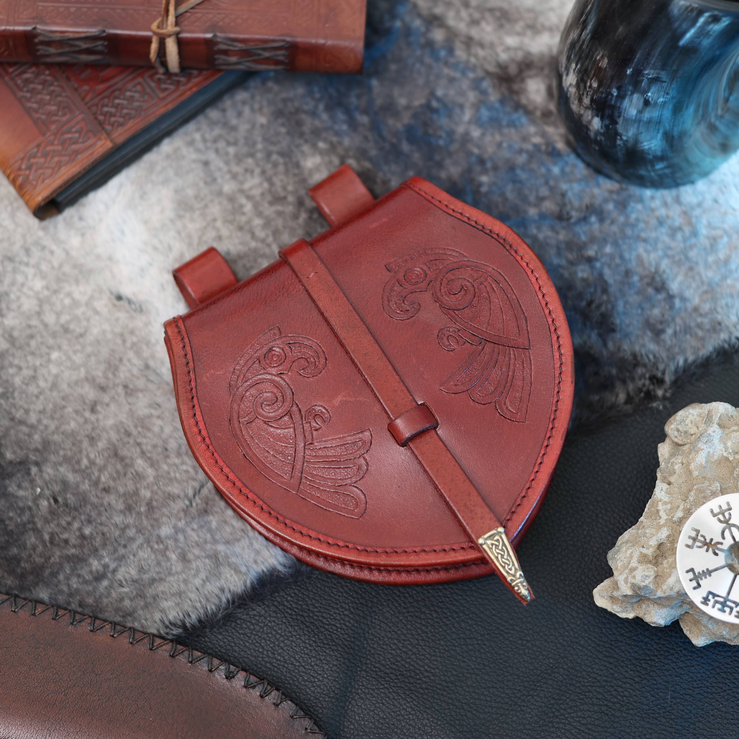 The "Huginn and Muninn" Viking Bag, featuring intricate embossing and a decorated clasp, rests on fur alongside a black leather item, a Norse mythology-inspired rune stone, and a journal.