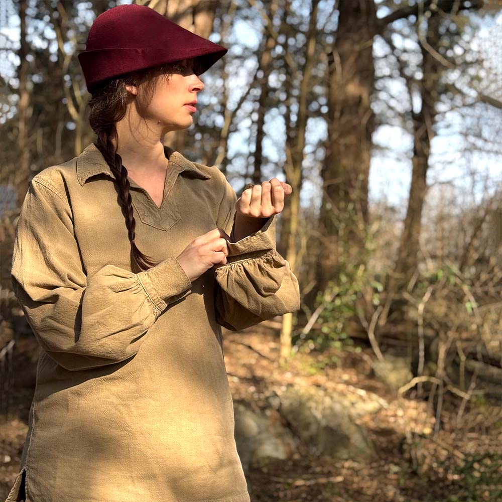 A person with a long braid and burgundy hat stands in a sunlit forest, wearing the Warrior Shirt – Medieval Strength & Style- Unisex in tan and adjusting a sleeve while looking to the left. Trees and rocks are visible in the background.