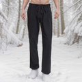 A barefoot man stands in the snow, wearing black Warrior Pants- Men's. Snow-covered trees fill the forest background, and his upper body is partially out of view.