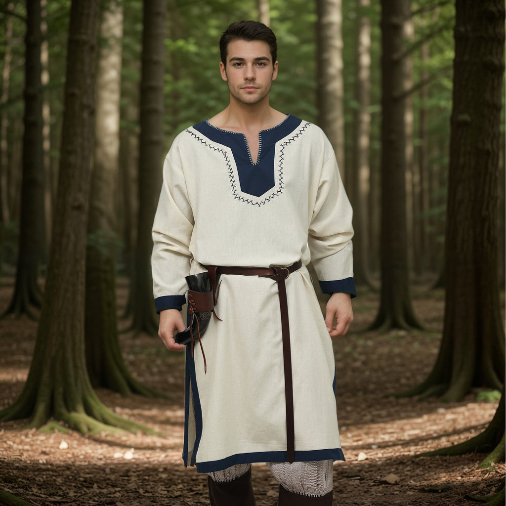 A man wears the Nordic/Viking Heritage Tunic for men—cream-colored with dark blue trim and a brown belt—as he stands in sunlight in a forest, holding a small pouch at his waist and looking calmly at the camera.
