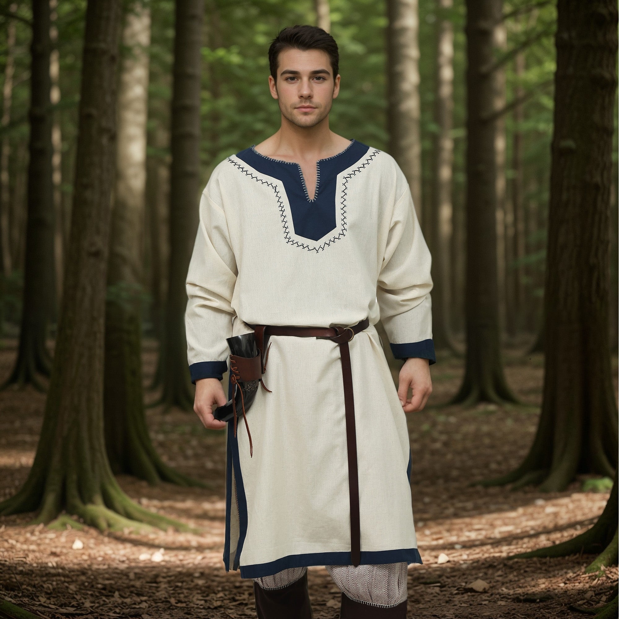 A man wears the Nordic/Viking Heritage Tunic for men—cream-colored with dark blue trim and a brown belt—as he stands in sunlight in a forest, holding a small pouch at his waist and looking calmly at the camera.