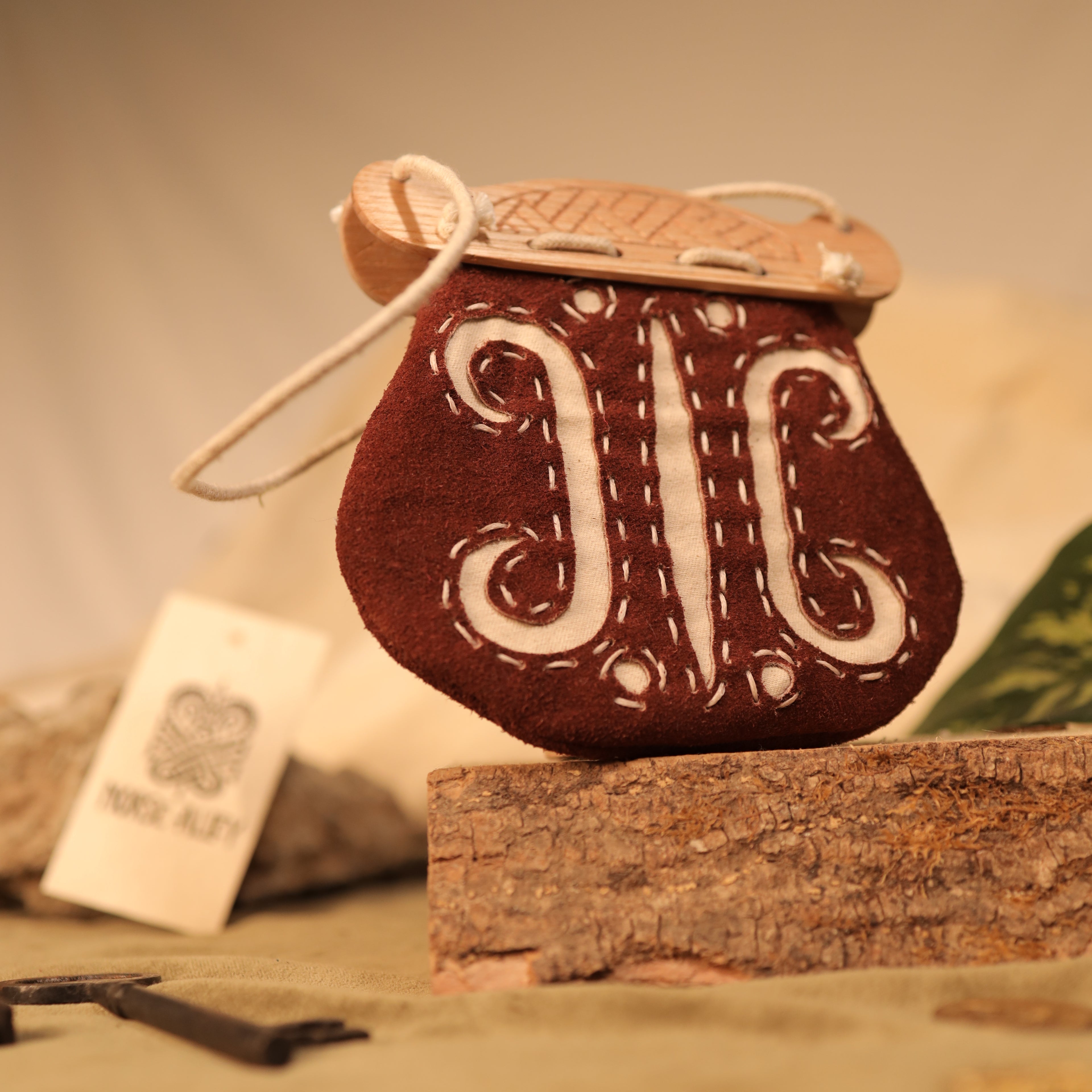 The "Gokstad" Coin Pouch is a handcrafted brown felt pouch with white embroidery and a wooden closure, displayed on wood with natural accents in the background.
