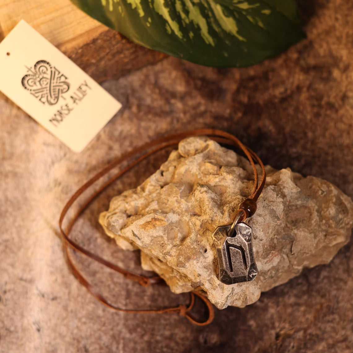 The Viking Rune Pendant Uruz, symbolizing strength and resilience, hangs from a leather cord on rough stone beside a Toke Alley tag with a Celtic knot. A green leaf is visible in the background.