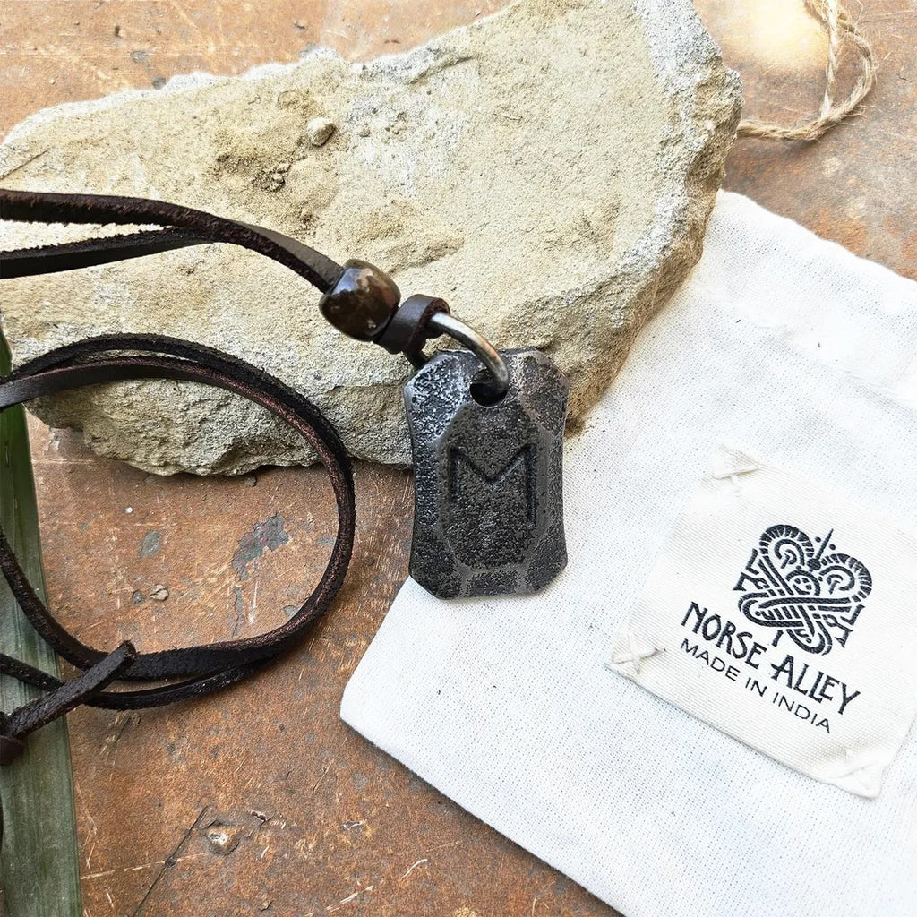 The Viking Rune Pendant Ehwaz, a dark metal Norse necklace with the M-shaped Ehwaz rune, hangs from a leather cord near a rough stone, next to a Norse Alley Made in India pouch with a Norse-style logo.