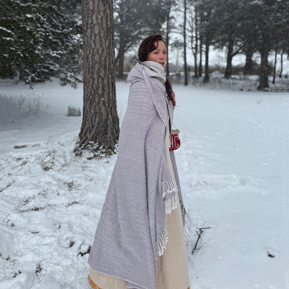 A woman stands in a snowy forest wearing Odin's Viking Cape - Unisex, a long light gray cape over a beige dress, with trees and snow-covered ground behind her. She looks back over her shoulder at the camera.