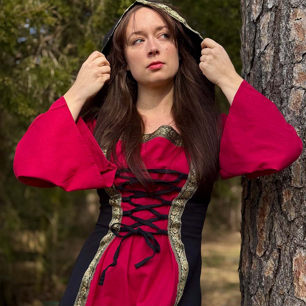 Wearing the Dark Majesty Medieval Dress—red and black with gold trim—a woman lifts her hood, standing by a tree in a forest, styled for a Renaissance fair.
