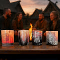 Four people in Viking-style clothing talk and laugh at sunset around a table set with decorated candles and Norse Alley Viking Drinking Horn Shot Glasses, with horn handicrafts nearby and a fire burning warmly in the background.