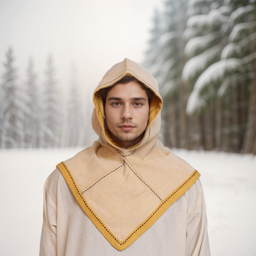 A person wearing a beige Reversible Viking Hood, Skjoldehamn, with yellow trim stands outdoors in a snowy evergreen forest, capturing the essence of medieval reenactment.