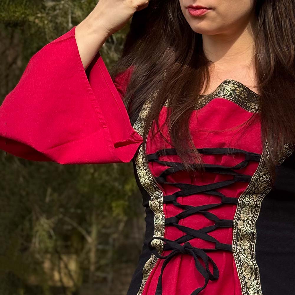 A woman wears the Dark Majesty Medieval Dress- Ladies in striking red and black with gold trim and a lace-up front—an ideal choice for Renaissance fairs or LARP. Her long brown hair cascades over her shoulders as she stands outdoors.