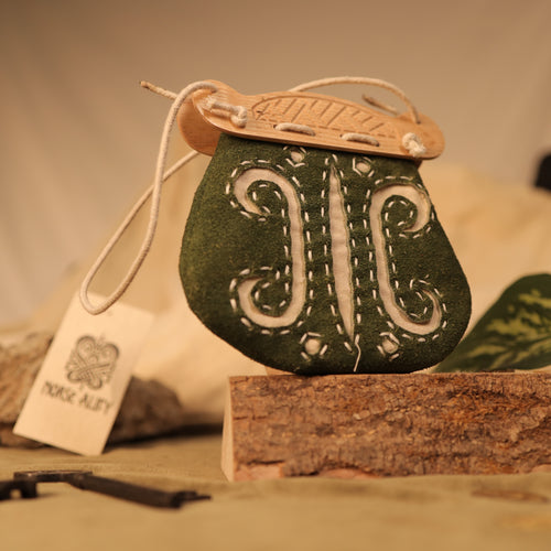 A small green leather "Gokstad" Coin Pouch with white stitching and a wooden top rests on bark. Beside this unique Viking pouch is a tag featuring a Viking knot design and the Nord Alley name.