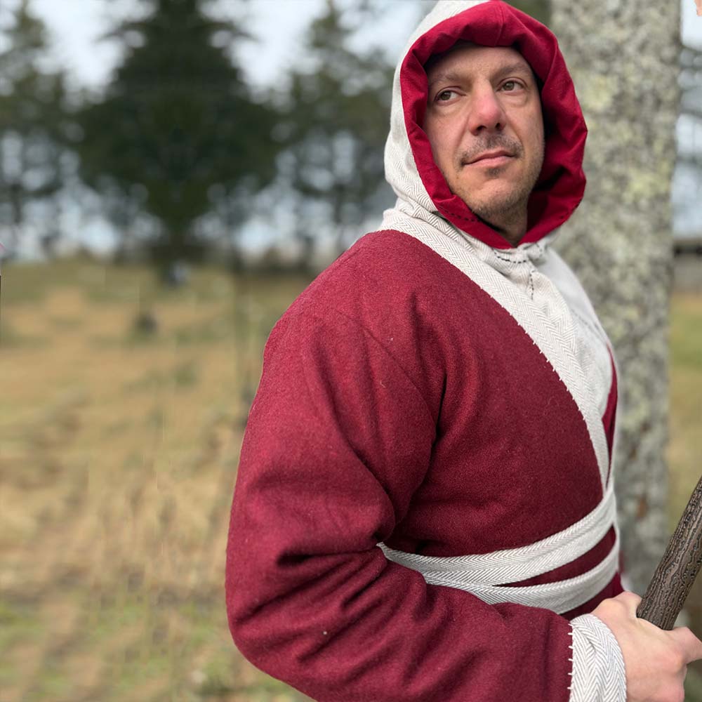 A person wearing a Viking Wool Coat - Men's in red and white stands outdoors near a tree, gazing thoughtfully into the distance. Tall evergreens blur in the background, evoking historical Viking attire.