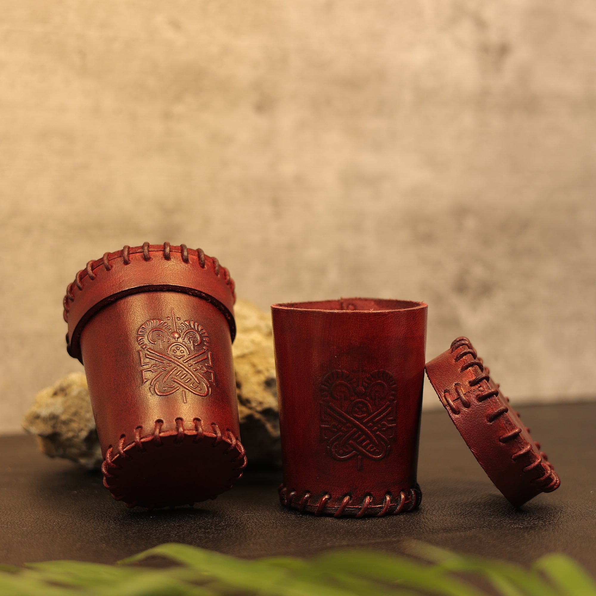 A Handcrafted Dark Red Leather Dice Cup with decorative stitching and embossed patterns is displayed—one upright, one on its side with the lid off—against a neutral background beside a rock and green plant.