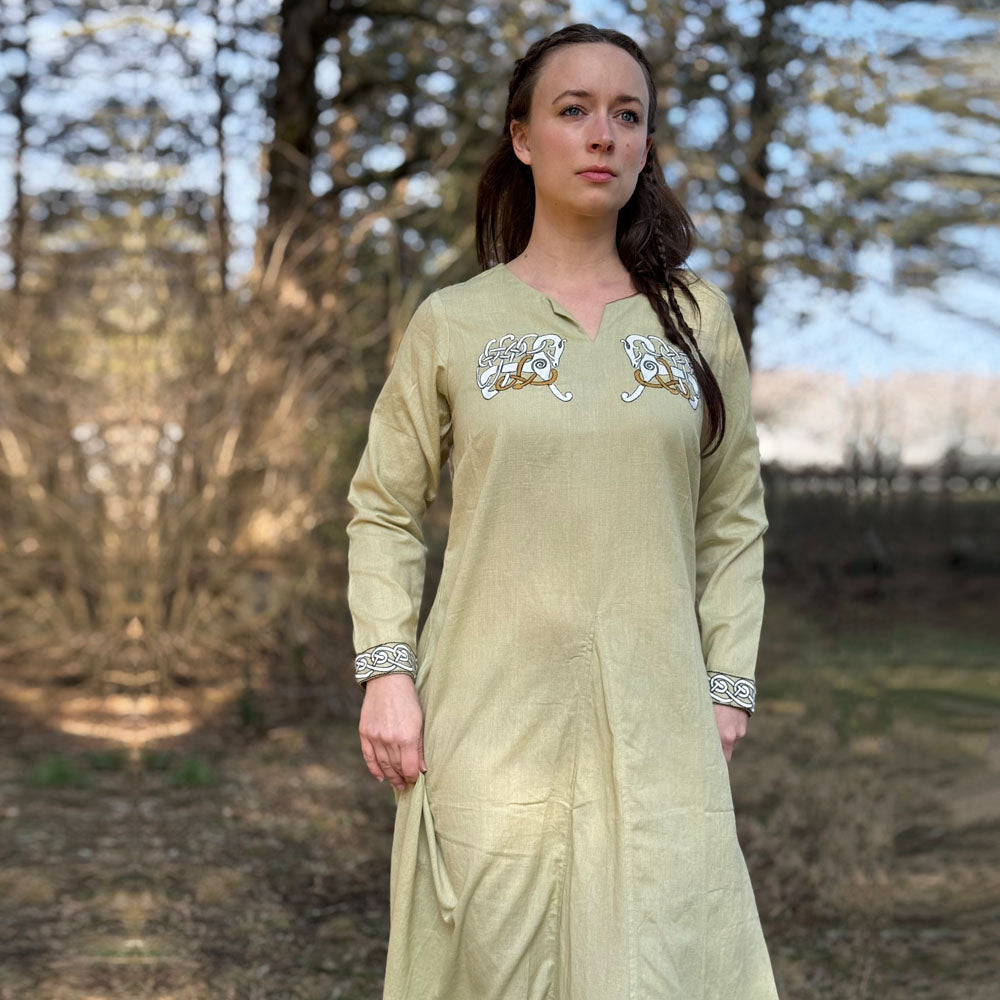 A woman stands in a sunlit wooded area wearing the Sleipnir Viking Dress—Ladies, a light beige cotton-linen gown featuring embroidered chest and cuffs. She gazes calmly ahead, surrounded by blurred trees.