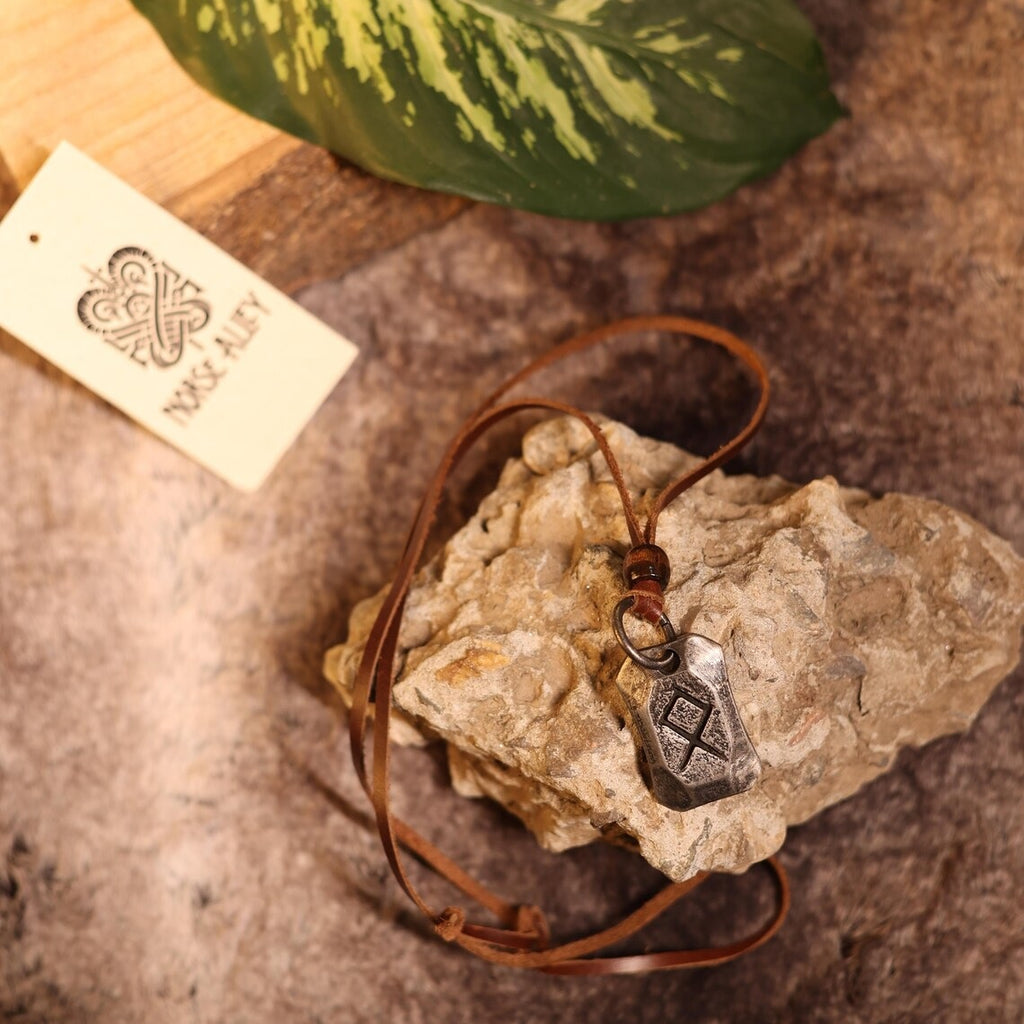 The Viking Rune Pendant Othila features an engraved rune on a rustic stone, attached to a brown leather cord. It’s displayed with a Norse Alley card on a textured surface, accented by a green leaf.