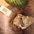 The Viking Rune Pendant Othila features an engraved rune on a rustic stone, attached to a brown leather cord. It’s displayed with a Norse Alley card on a textured surface, accented by a green leaf.