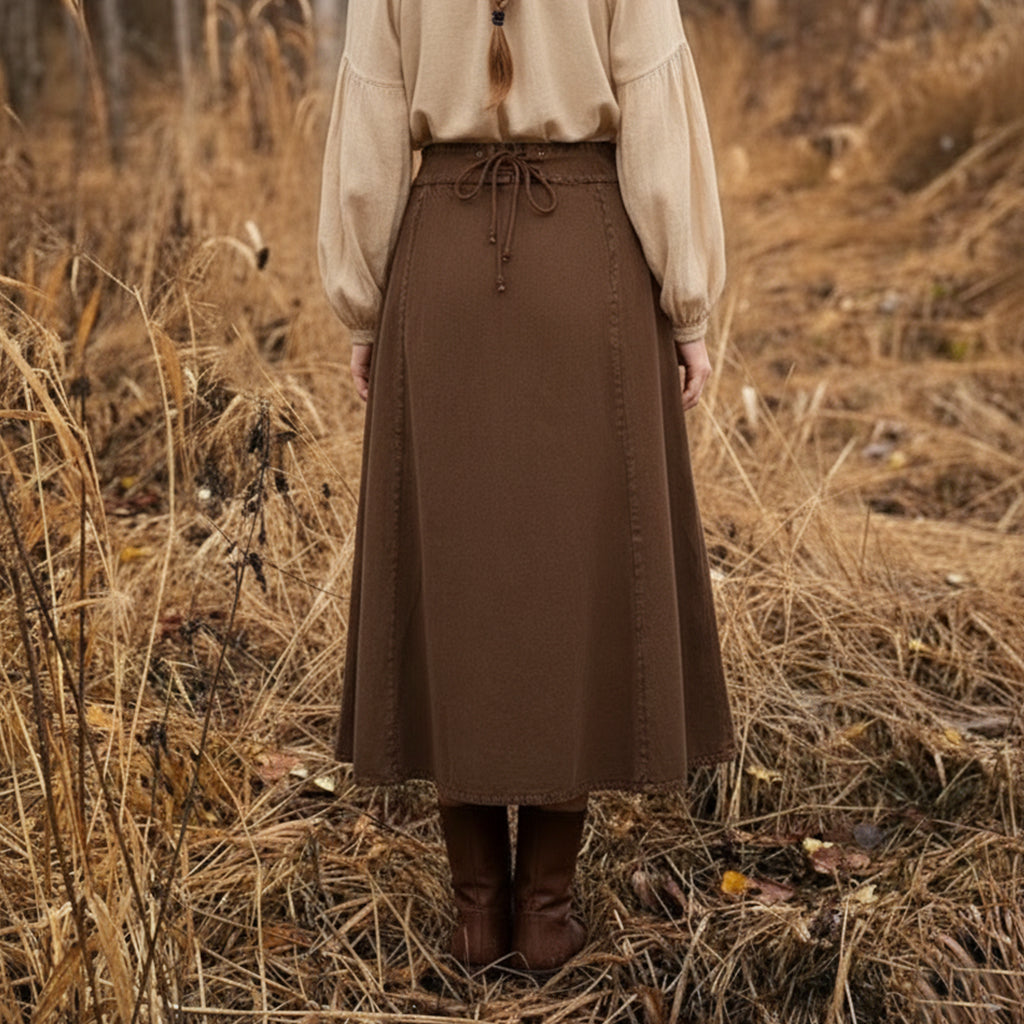 A person stands in a field of dry tall grass, seen from behind, wearing a beige long-sleeve blouse and the Medieval Skirt- Ladies—a brown midi skirt with waist tie—paired with brown boots for a Renaissance-inspired look.