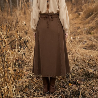 A person stands in a field of dry tall grass, seen from behind, wearing a beige long-sleeve blouse and the Medieval Skirt- Ladies—a brown midi skirt with waist tie—paired with brown boots for a Renaissance-inspired look.