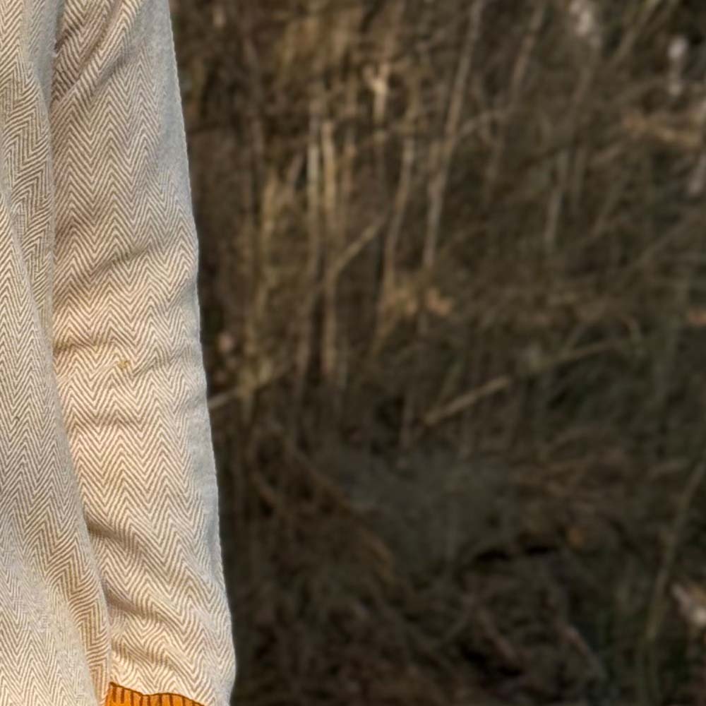 A close-up of a person wearing the Nordic Runeweave Viking Dress—cream herringbone fabric with hand embroidery and orange cuff—stands outdoors before blurred dry grass, highlighting Norse craftsmanship.
