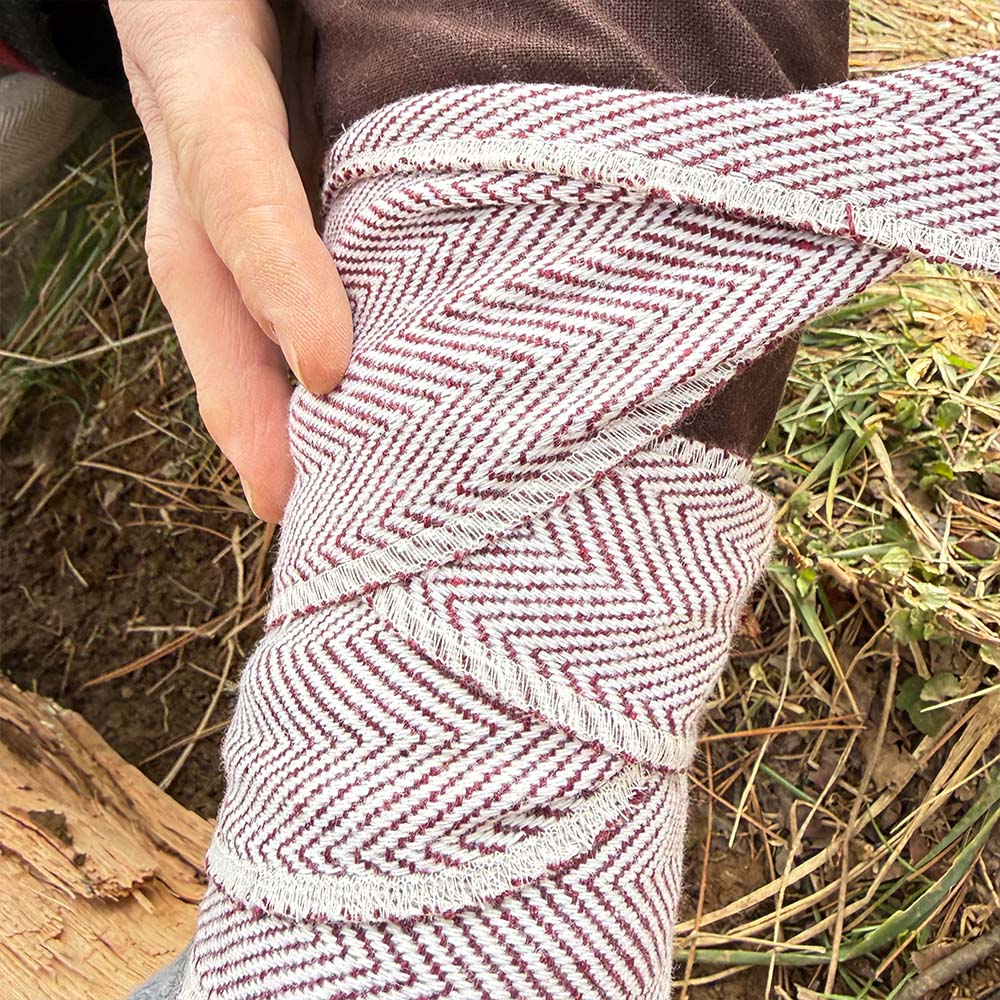 A hand wraps Herringbone Viking Leg Wraps (Winingas), featuring a chevron design, around a person's lower leg outdoors near grass and wood.