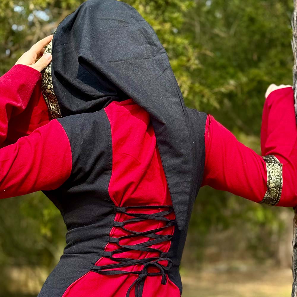 A person stands outdoors with their back to the camera, wearing the Dark Majesty Medieval Dress- Ladies in red and black with gold trim and a hood. This LARP-ready dress features lace-up detailing on the back, framed by green trees in the background.