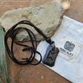 The Viking Rune Pendant Perth necklace, featuring a Perthro-like symbol, lies on a stone next to a Norse Alley bag made in India, resting on a rustic surface with a green leaf close by.