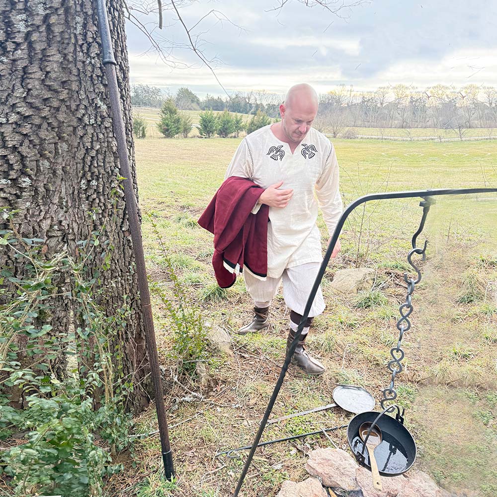 A man in Odin's Raven Tunic with dark embroidery walks near a tree, holding a maroon cloth. An iron tripod supports a pot over a stone fire pit in a grassy field beneath cloudy skies.