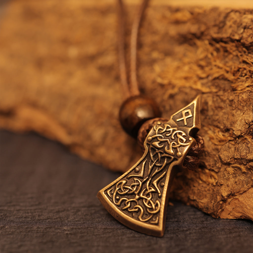 The Viking Axe Brass Pendant showcases intricate knotwork and a rune symbol, highlighted by a wooden bead on the cord. Resting on a coarse brown surface, this Norse jewelry makes a striking addition to any collection.
